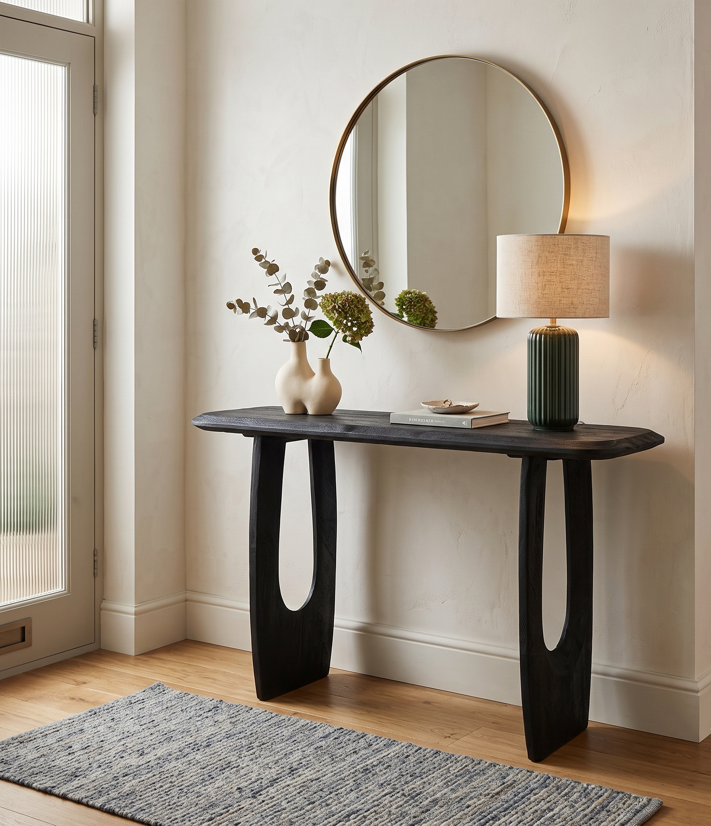 A dark wood console table with sculptural cutout legs in a styled entryway. A round wall mirror, table lamp, two small vases with dried flowers, and a stack of books sit on the tabletop.