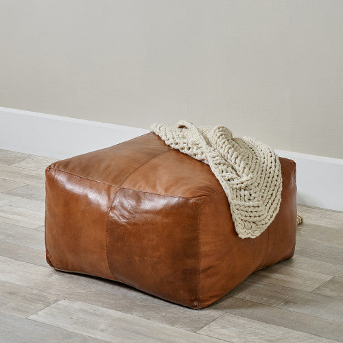 Brown leather ottoman with a white knitted blanket on a wooden floor.