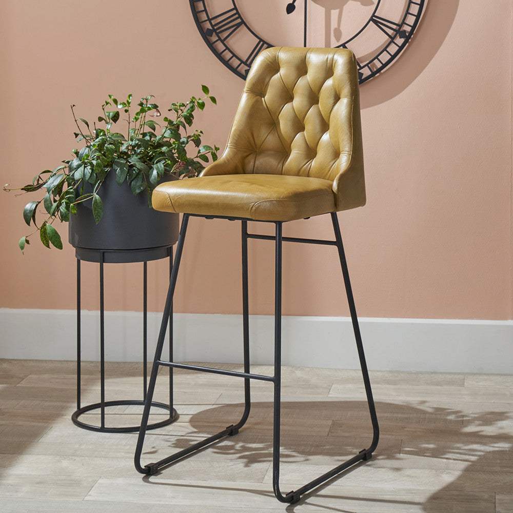 Yellow bar stool with black metal legs next to a potted plant against a beige wall with a clock.