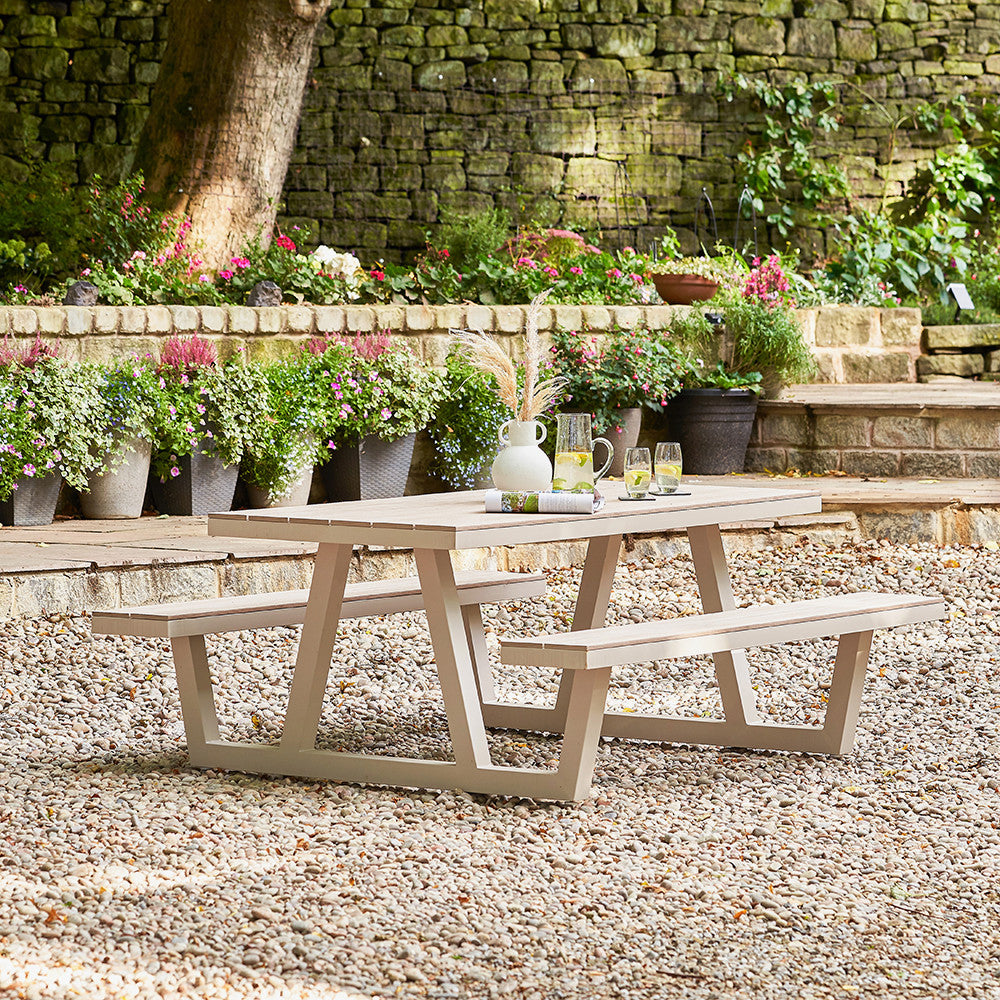 Outdoor wooden picnic table with benches in a garden setting with potted plants and a stone wall.