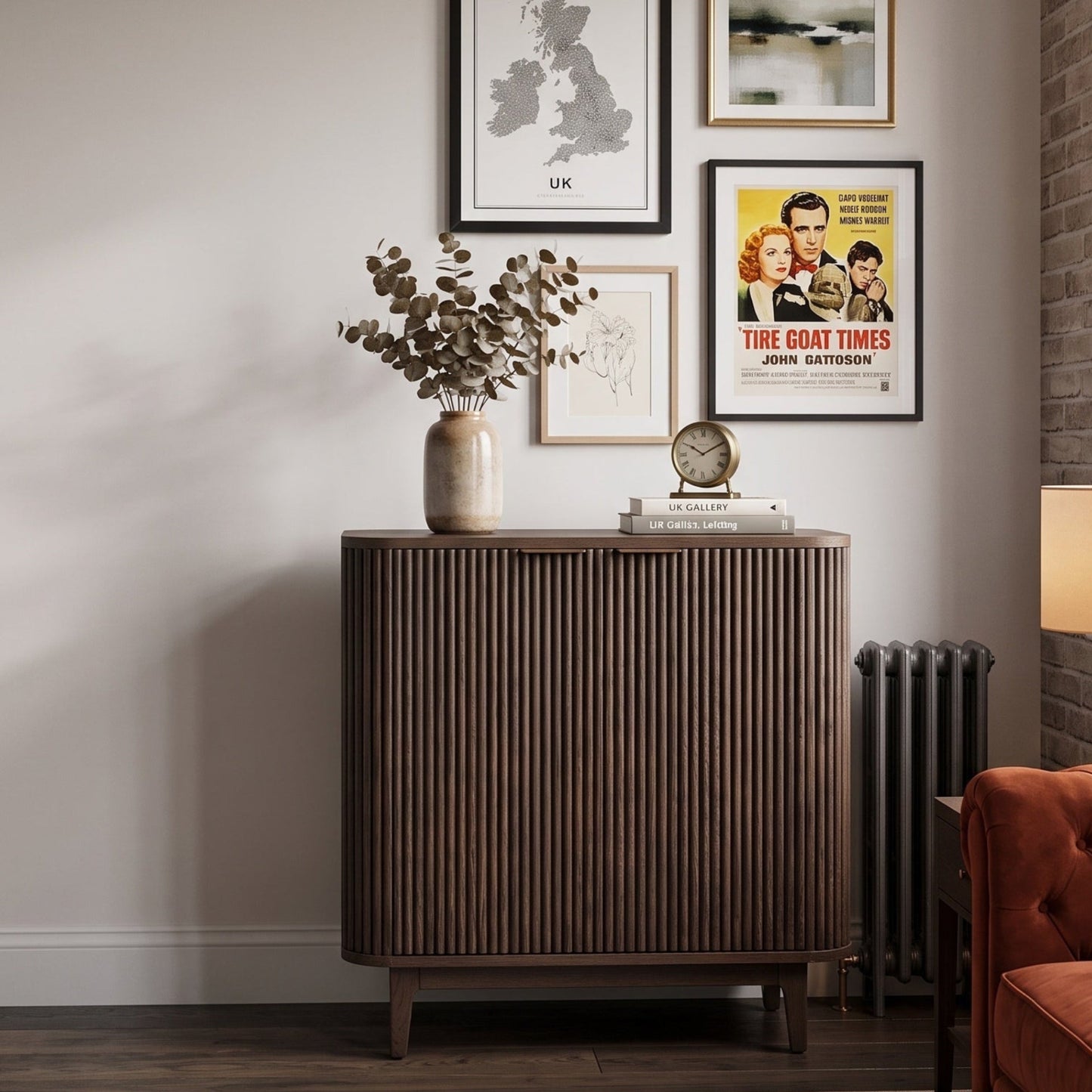 A tall ribbed wood cabinet in a warm medium brown tone stands against a light gray wall in a styled room. Framed wall art hangs above it, with a vase of branches on top, and part of an orange-brown sofa and a window are visible to the right.