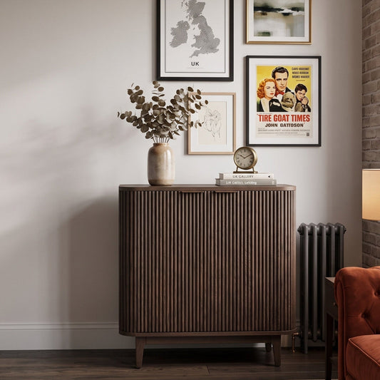A tall ribbed wood cabinet in a warm medium brown tone stands against a light gray wall in a styled room. Framed wall art hangs above it, with a vase of branches on top, and part of an orange-brown sofa and a window are visible to the right.