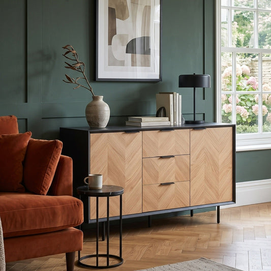 A modern wooden sideboard cabinet with a chevron pattern on the drawer fronts, set in a styled living room with a dark green paneled wall, an orange-brown armchair, and a window.