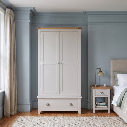 A white double-door wardrobe with a light wood top and silver knobs, positioned in a styled bedroom with blue walls, a window, and a bed.