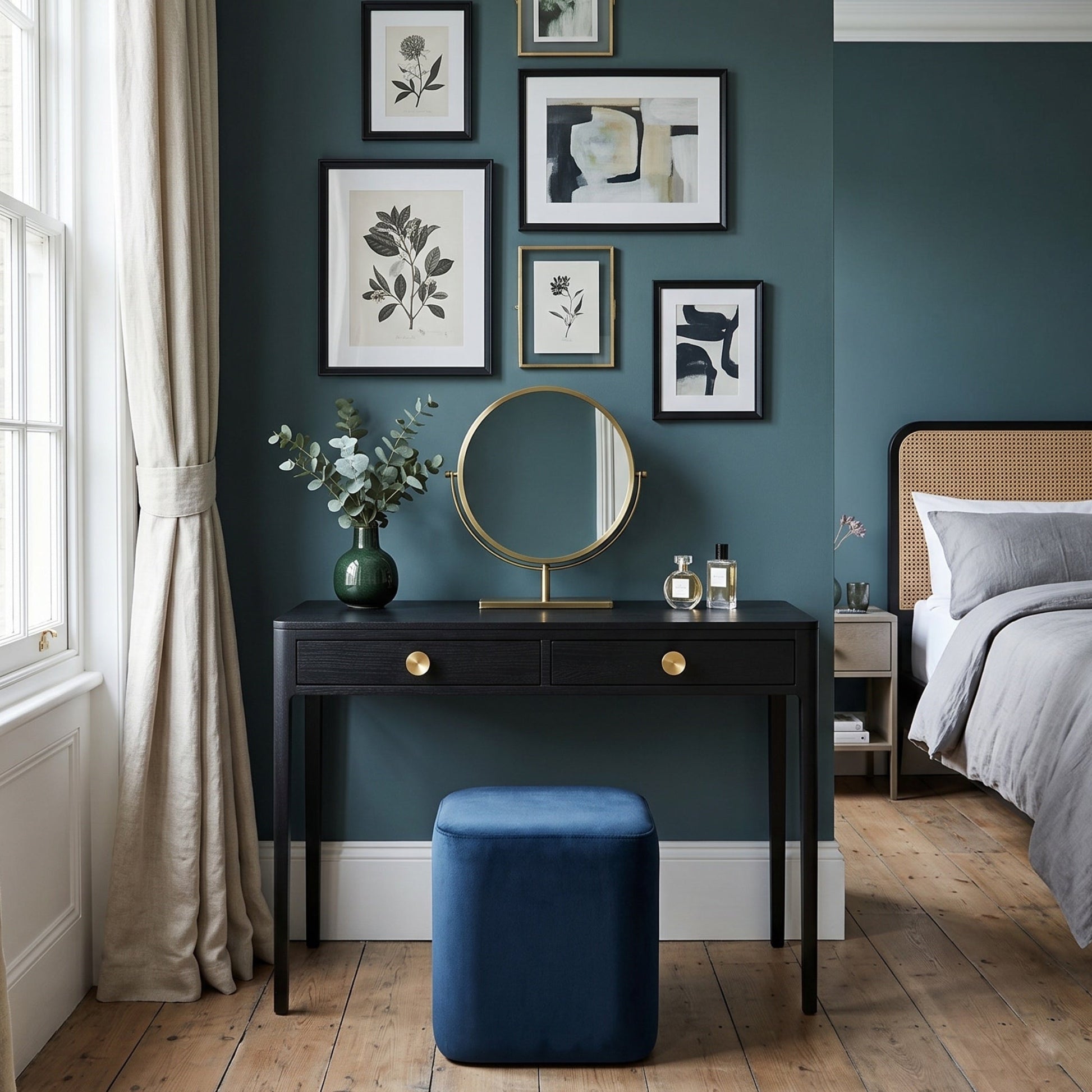 A styled bedroom interior featuring a dark teal wall with framed botanical and abstract art prints, a black vanity table with a round gold-framed mirror, a blue velvet stool, a window with beige curtains, and a bed with grey bedding.