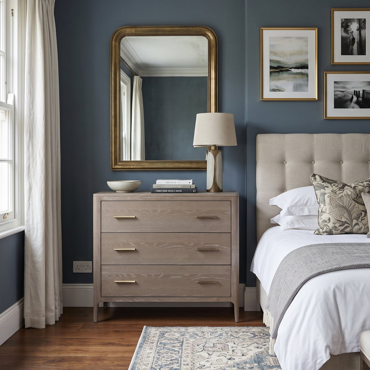 A styled bedroom interior featuring a light wood three-drawer dresser with a large gold-framed mirror, a table lamp, and a tufted beige bed with white bedding, set against dark blue walls.