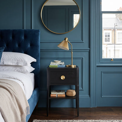 A styled bedroom interior featuring a deep blue tufted bed headboard, white bedding, a black nightstand with brass hardware, a gold desk lamp, a round gold-framed mirror, and a blue paneled wall.