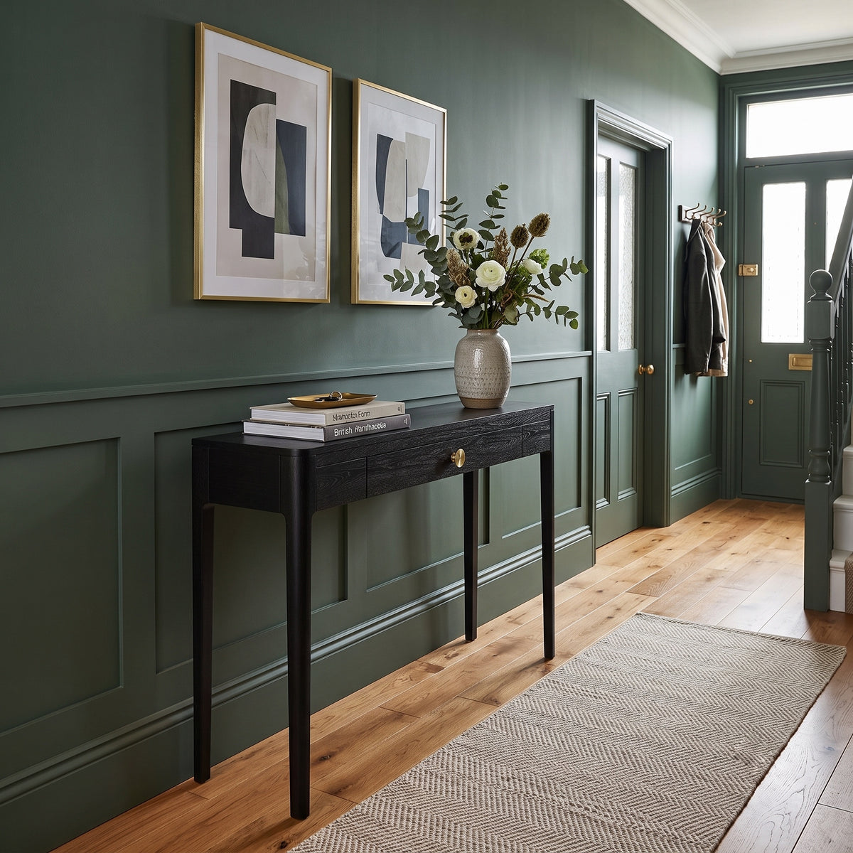A dark wood console table with a single drawer and brass knob, styled with books and a ceramic vase of greenery, set against a dark green paneled wall in a beautifully decorated entryway.