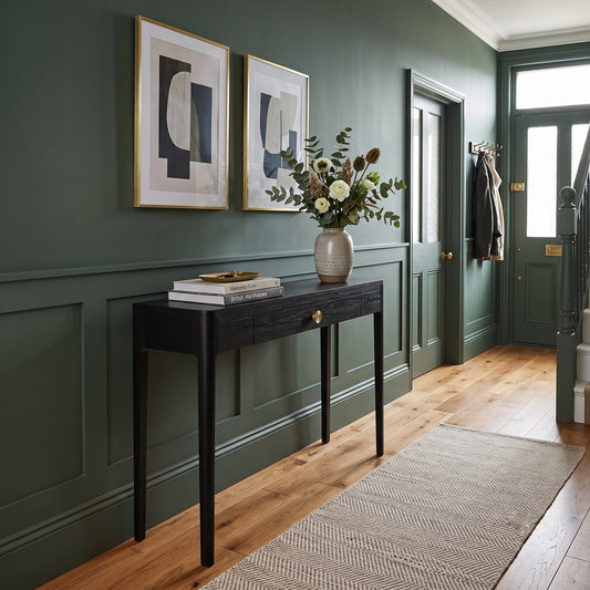 A dark wood console table with a single drawer and brass knob, styled with books and a ceramic vase of greenery, set against a dark green paneled wall in a beautifully decorated entryway.