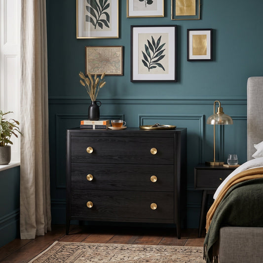 A dark wood chest of drawers with round gold knobs stands in a styled bedroom with a teal wall, framed botanical prints, a bedside lamp, and part of a bed with green and gold bedding.
