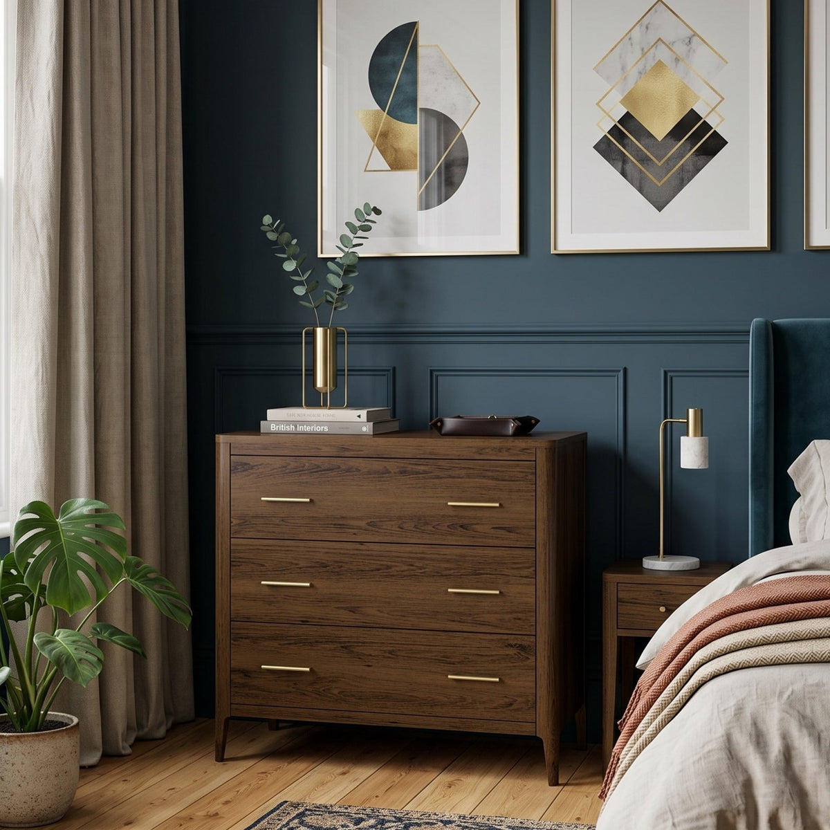 A styled bedroom interior featuring a dark wood three-drawer dresser, a bed with neutral linens, framed geometric wall art, and a deep navy blue wall. The dresser has gold-toned handles and sits next to a window with beige curtains.