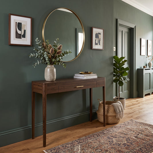 A styled interior scene with a dark wood console table against a deep green wall, topped with a ceramic vase of dried flowers, books, and a round wall mirror. Framed wall art hangs on the wall, a potted plant stands near a doorway, and a woven rug covers the wood floor.