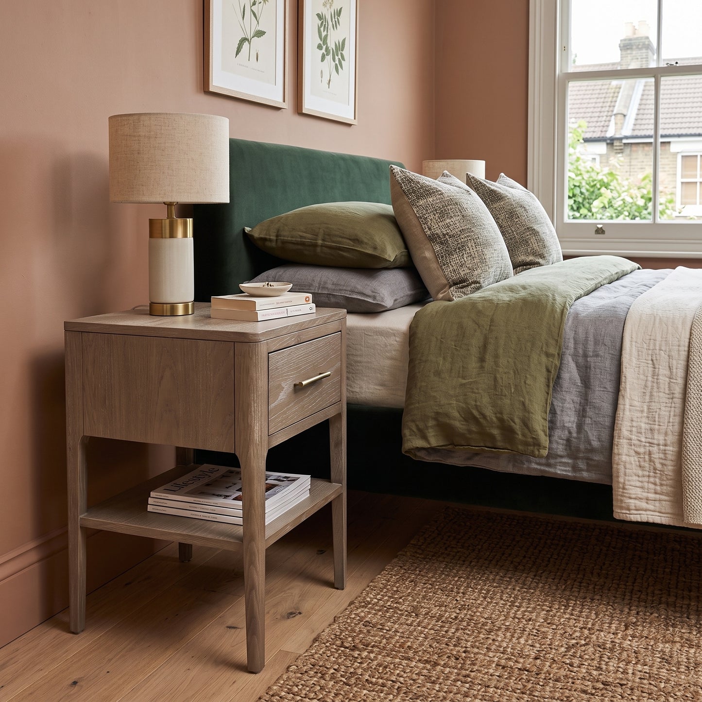 A rustic light oak nightstand in a styled bedroom setting, featuring a beige drum lamp, stacked books, and a small magazine on its open shelf.