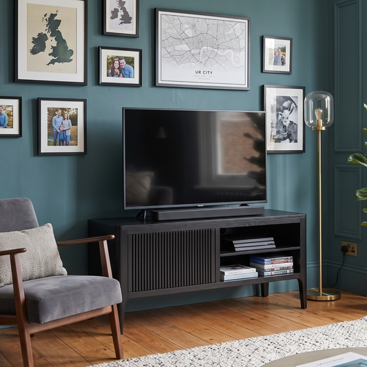 A styled living room corner with a dark gray armchair, a black TV stand holding a flat-screen television, a teal wall with framed artwork, a potted fiddle-leaf fig plant, and a round coffee table in the foreground.