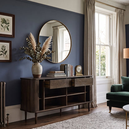 A styled living room corner featuring a dark wood console table beneath a round wall mirror, set against a deep blue wall with framed botanical prints. A green sofa, beige curtains, and a textured rug are visible in the room.