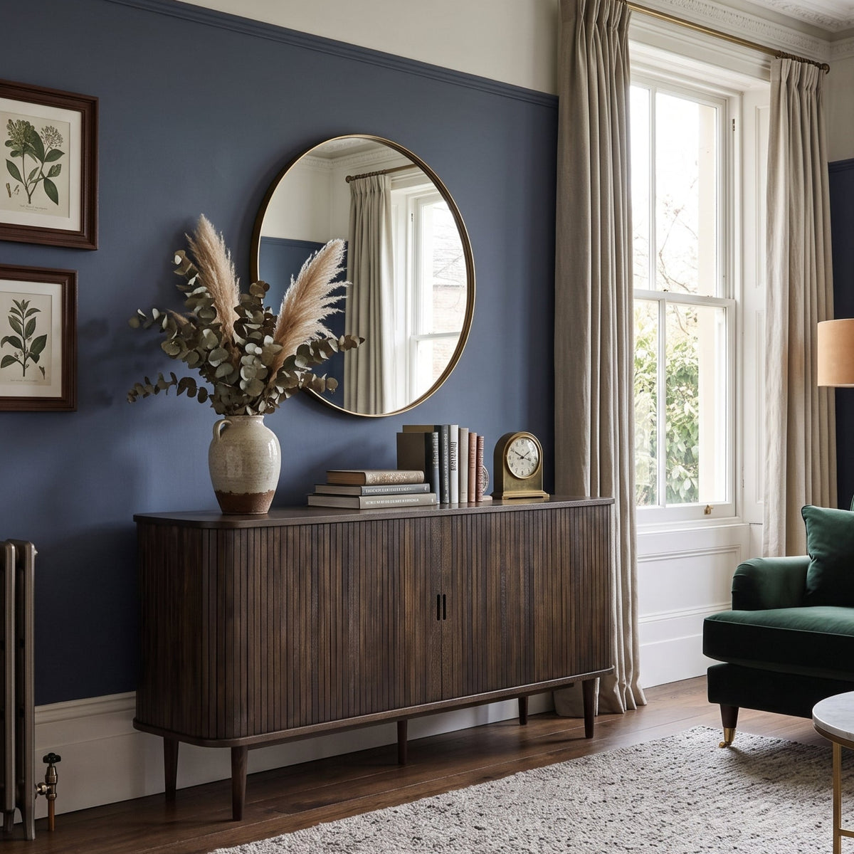 A styled living room interior featuring a mid-century modern dark wood credenza under a round wall mirror, set against a deep blue accent wall with framed botanical prints. A green velvet sofa, a round coffee table, and a textured rug are visible in the foreground.