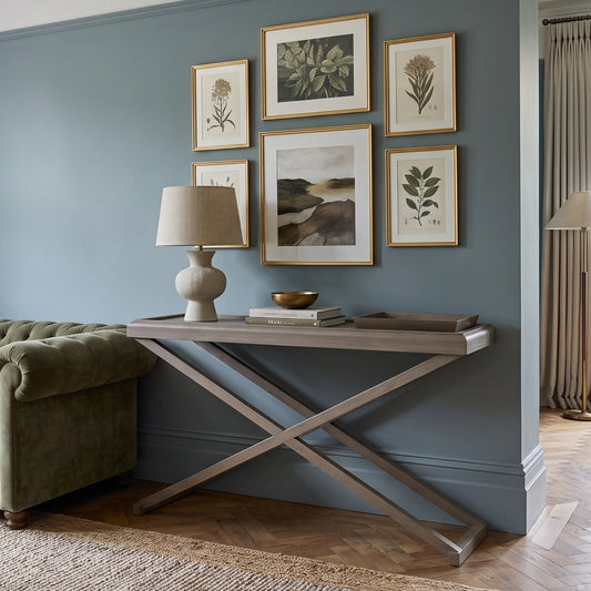 A styled interior room scene showing a blue-gray accent wall with six framed botanical art prints, a light wood console table with an X base, a cream table lamp, a green tufted sofa, and a window with curtains in the background.