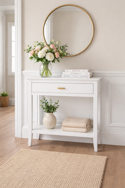 White console table with a round mirror, flowers, and books in a room with wooden flooring and a rug.