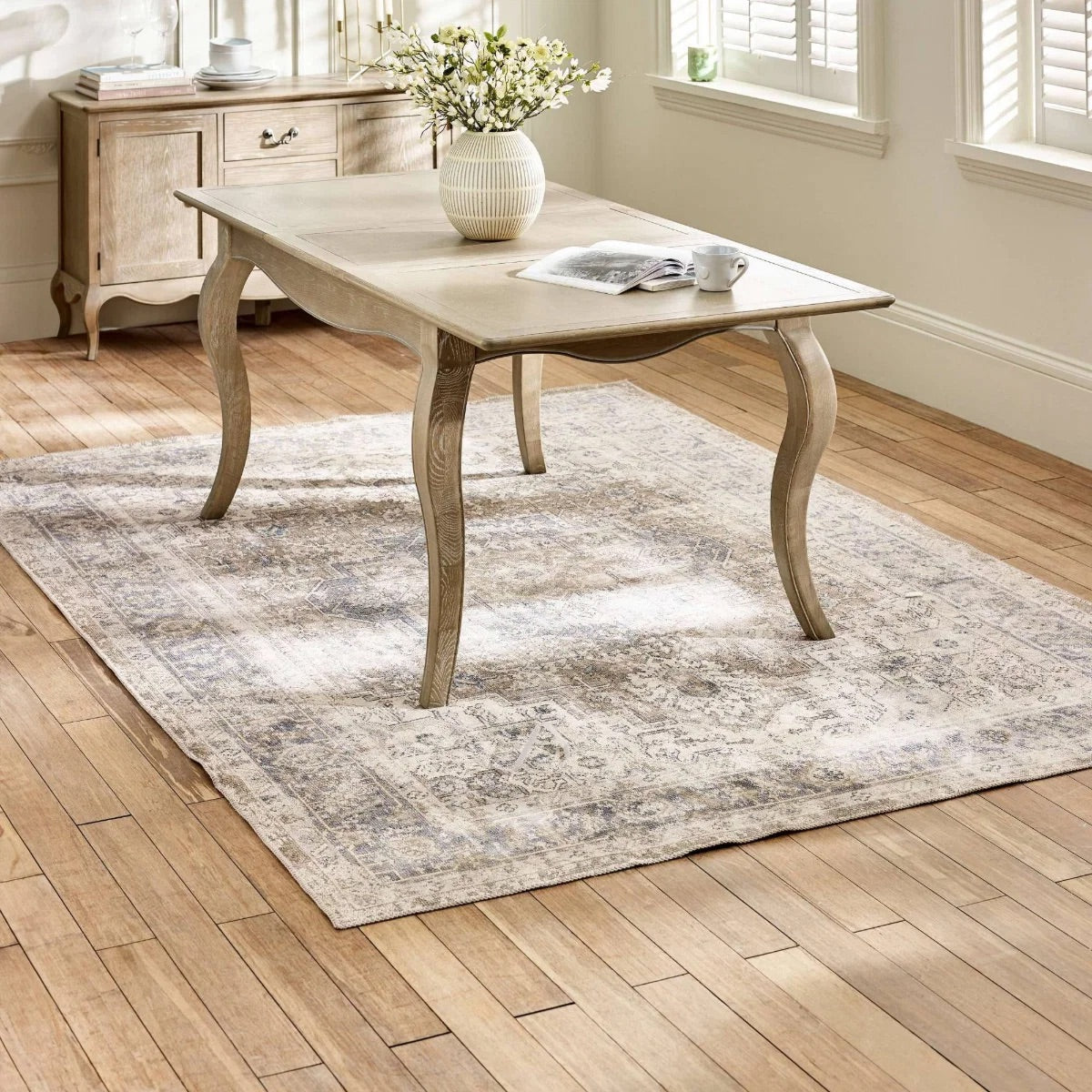 Wooden dining table with a vase of flowers and a newspaper on a patterned rug in a room with wooden flooring.
