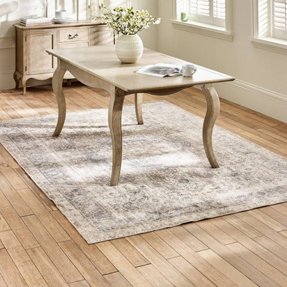 Wooden dining table with a vase of flowers and a newspaper on a patterned rug in a room with wooden flooring.