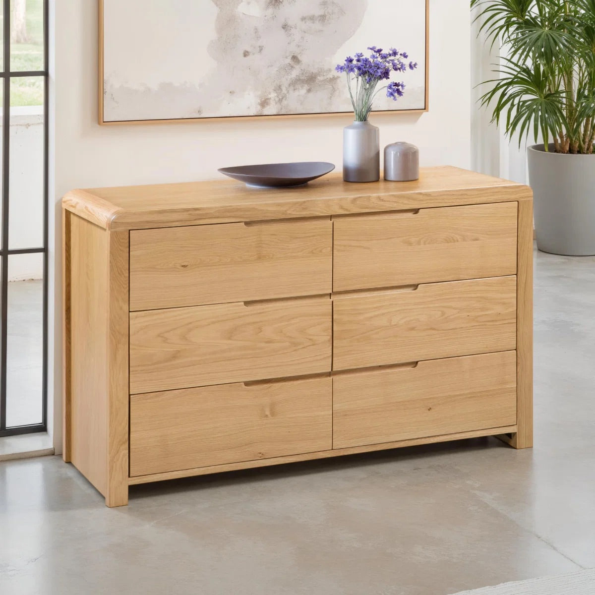Wooden dresser with decorative items in a room with a plant and framed artwork.