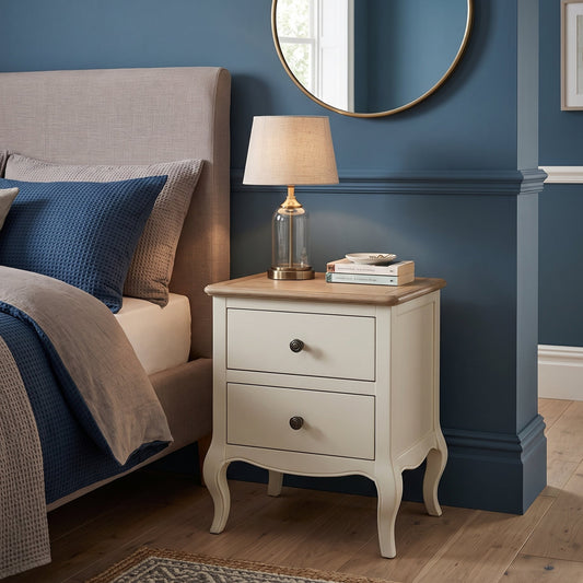 A classic two-drawer nightstand in cream with a light wood top, styled in a bedroom next to a bed with blue bedding and a table lamp.