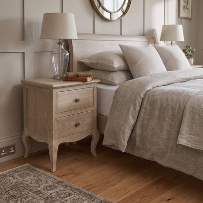 A light natural wood nightstand with two drawers and curved legs stands beside a bed in a softly lit bedroom. The nightstand has a light beige fabric lampshade on a clear glass base and a small stack of books on top.
