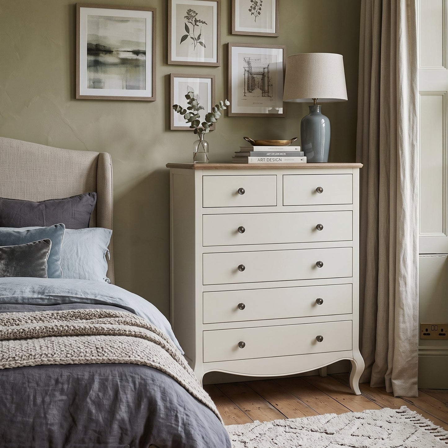 An off-white wooden five-drawer chest of drawers styled in a cozy bedroom next to a bed with gray bedding, set against a sage green wall with framed art and a window with beige curtains.
