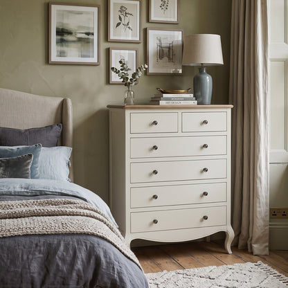 An off-white wooden five-drawer chest of drawers styled in a cozy bedroom next to a bed with gray bedding, set against a sage green wall with framed art and a window with beige curtains.
