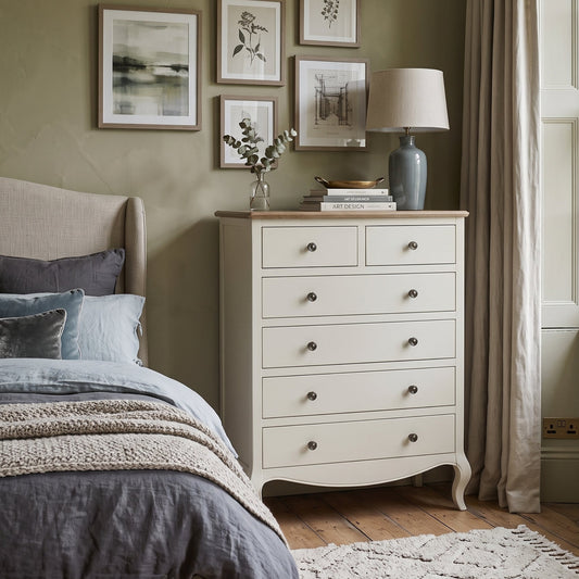 An off-white wooden five-drawer chest of drawers styled in a cozy bedroom next to a bed with gray bedding, set against a sage green wall with framed art and a window with beige curtains.