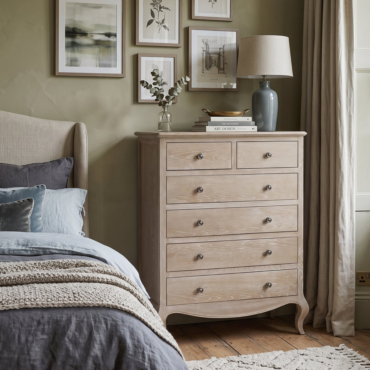 A light natural wood five-drawer dresser in a styled bedroom setting, positioned next to a bed with gray and blue bedding. The dresser features curved legs, a top surface with two small drawers and one larger compartment, and small round metal knobs.