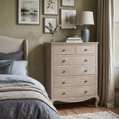 A light natural wood five-drawer dresser in a styled bedroom setting, positioned next to a bed with gray and blue bedding. The dresser features curved legs, a top surface with two small drawers and one larger compartment, and small round metal knobs.