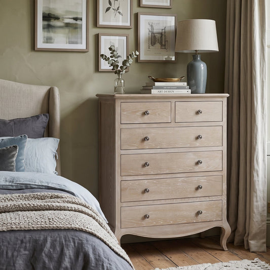 A light natural wood five-drawer dresser in a styled bedroom setting, positioned next to a bed with gray and blue bedding. The dresser features curved legs, a top surface with two small drawers and one larger compartment, and small round metal knobs.
