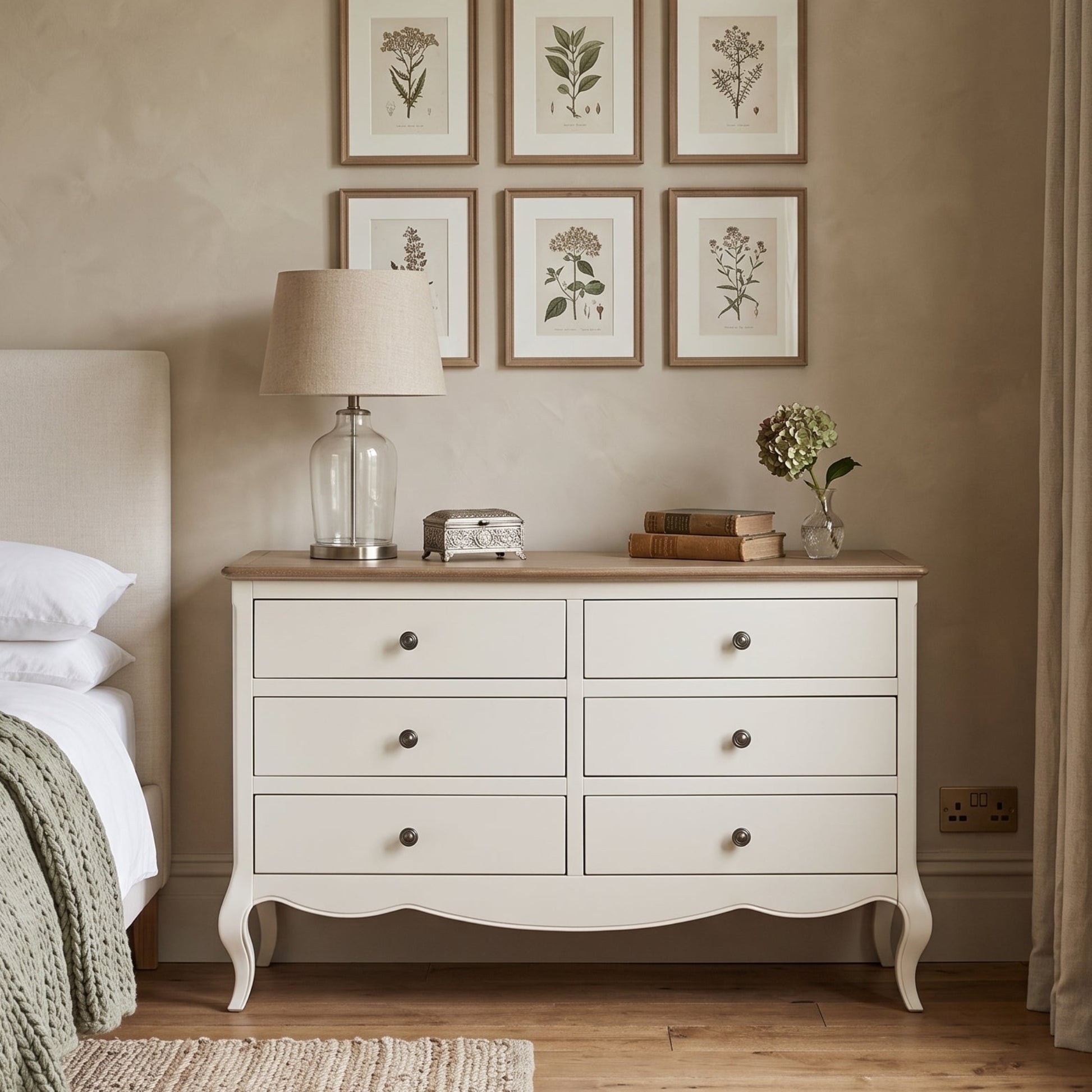 A white six-drawer dresser with a light wood top stands in a styled bedroom next to a bed. The dresser is decorated with a glass lamp, a small clock, books, and a vase of flowers.
