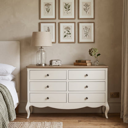 A white six-drawer dresser with a light wood top stands in a styled bedroom next to a bed. The dresser is decorated with a glass lamp, a small clock, books, and a vase of flowers.