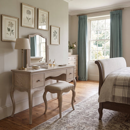 A light beige wooden vanity table with a matching upholstered stool, a wall mirror, and a table lamp, styled in a classic bedroom setting with a window, teal curtains, and botanical wall art.