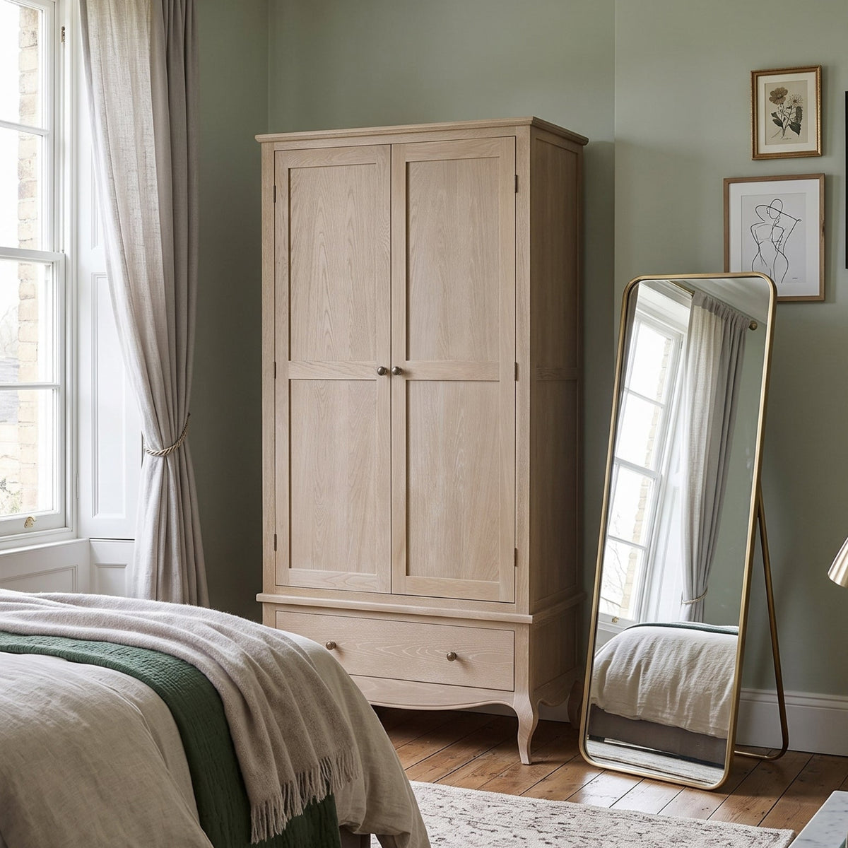 A light natural wood double-door wardrobe with curved legs and a single drawer, styled in a classic bedroom with sage green walls, a full-length floor mirror, and a bed.