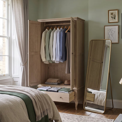 A styled bedroom interior featuring a tall light wood wardrobe with open doors and hanging shirts, a full-length floor mirror, a bed with neutral bedding, framed wall art, and soft sage green walls.