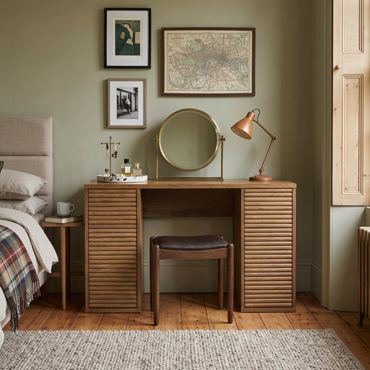 A wooden vanity desk with louvered side panels and a matching stool in a styled bedroom. A copper desk lamp, small mirror, and decorative items sit on the desk, with framed wall art above and a bed visible on the left.