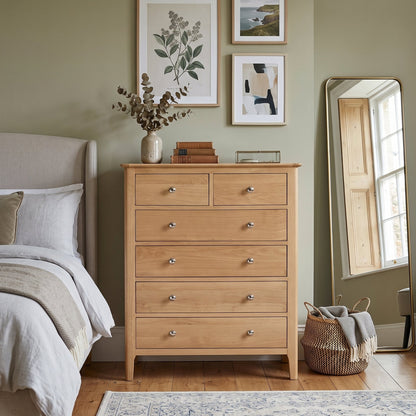 A light oak wood five-drawer chest of drawers in a styled bedroom setting, flanked by a bed and an upright mirror against a sage green wall.