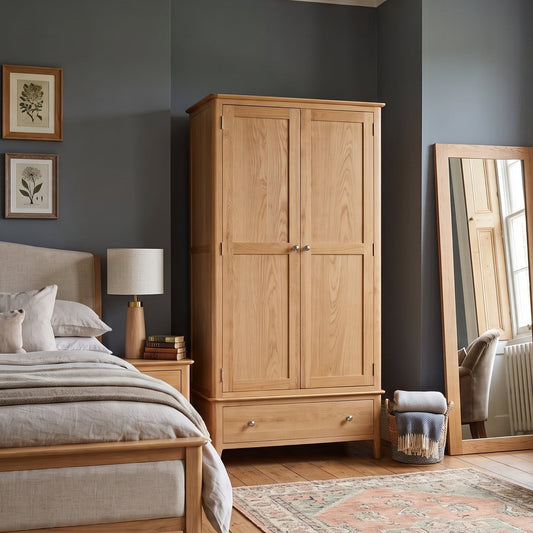 A styled bedroom featuring a light natural oak double-door wardrobe, a matching wooden bed frame, a large floor mirror, and a patterned area rug against dark slate-gray walls.