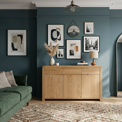 A styled living room with dark teal walls, a green sofa, a light wood sideboard, framed wall art, a large floor mirror, and potted plants.