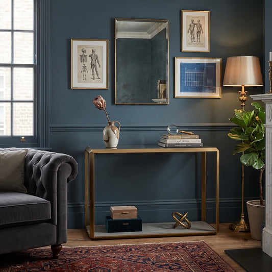 A styled interior scene with a gold-framed console table against a dark blue wall, surrounded by framed art, a mirror, a lamp, a plant, a sofa, and a rug.