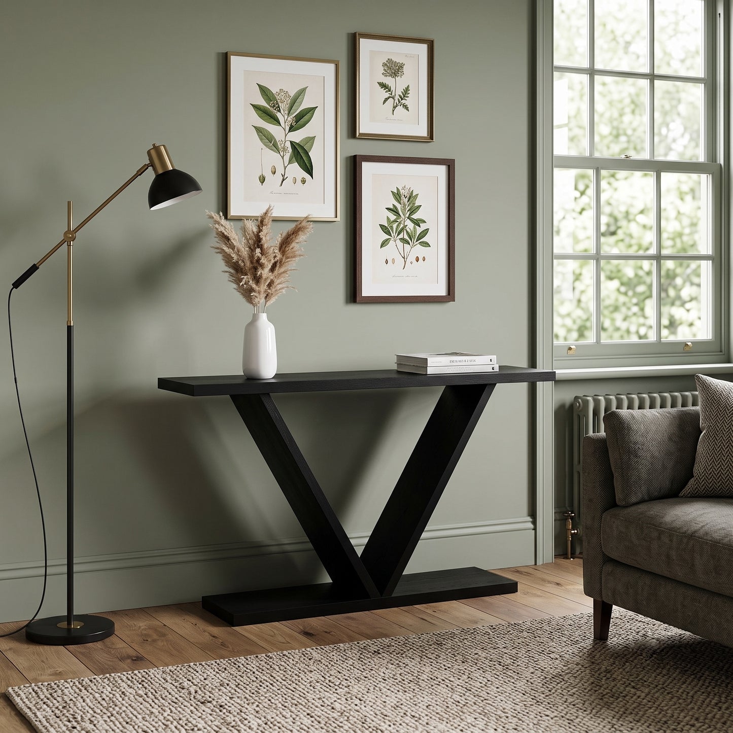 A modern black console table with a V-shaped metal base in a styled living room setting, featuring a white vase with dried grass and a book on top, next to a black floor lamp and a gray armchair.