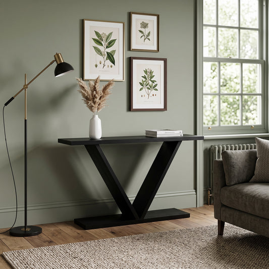 A modern black console table with a V-shaped metal base in a styled living room setting, featuring a white vase with dried grass and a book on top, next to a black floor lamp and a gray armchair.