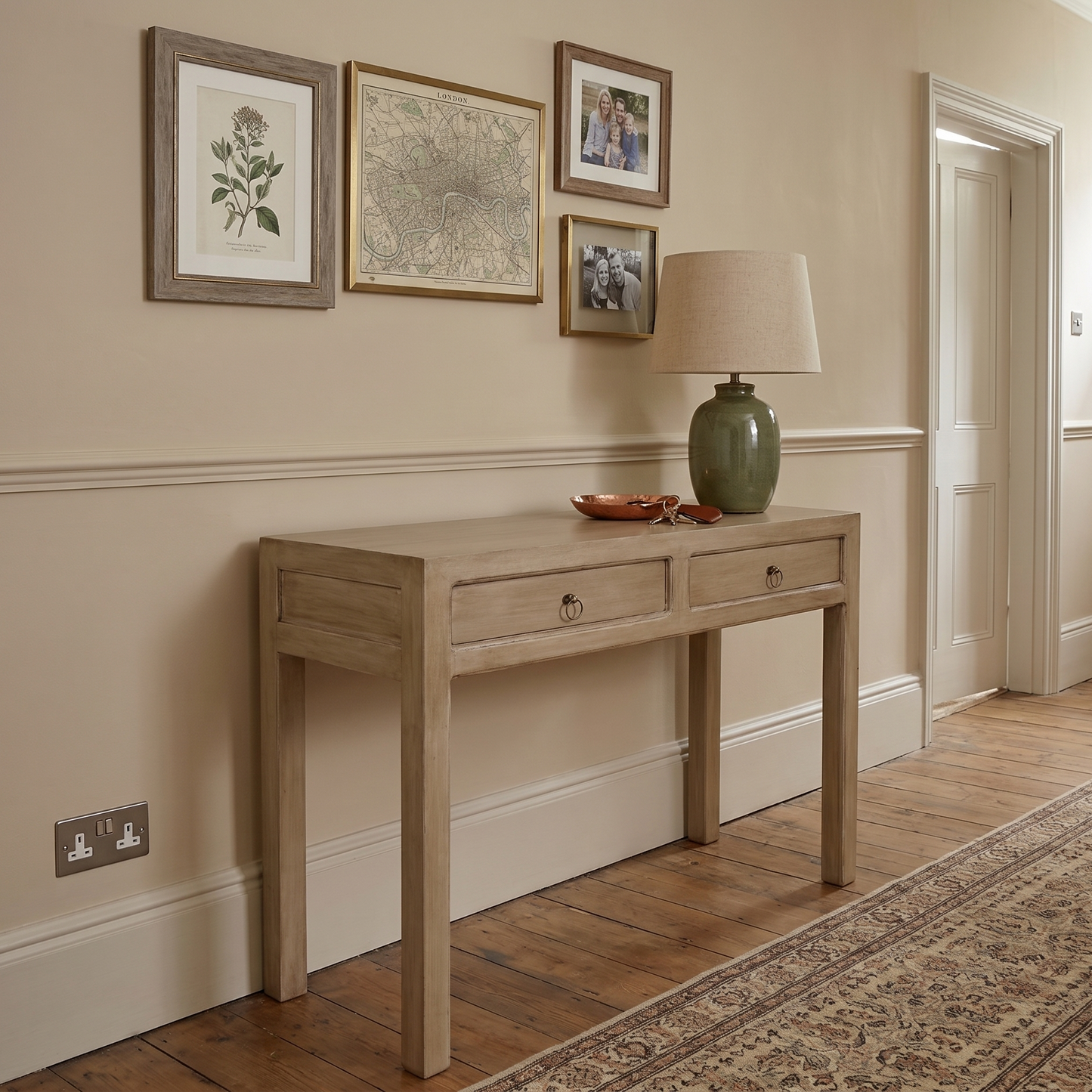 A light natural wood console table with two drawers and small round metal knobs, styled in a traditional hallway with a green ceramic table lamp, framed botanical prints, and a patterned area rug.