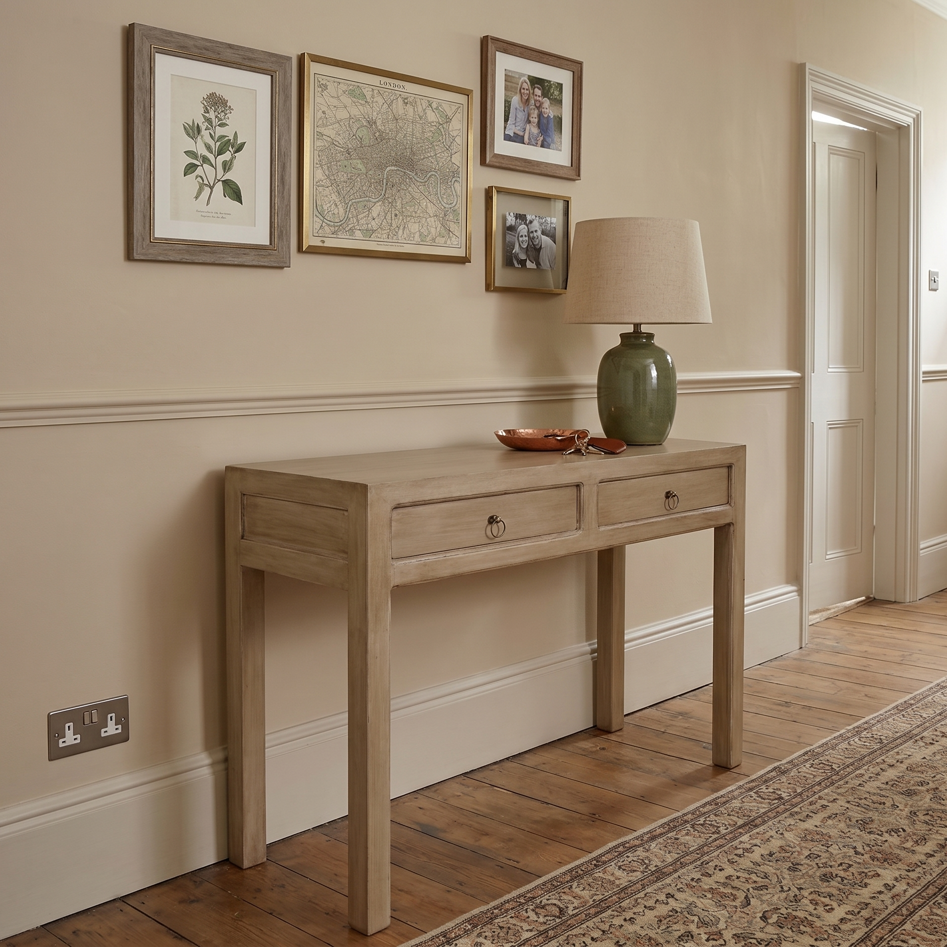 A light natural wood console table with two drawers and small round metal knobs, styled in a traditional hallway with a green ceramic table lamp, framed botanical prints, and a patterned area rug.
