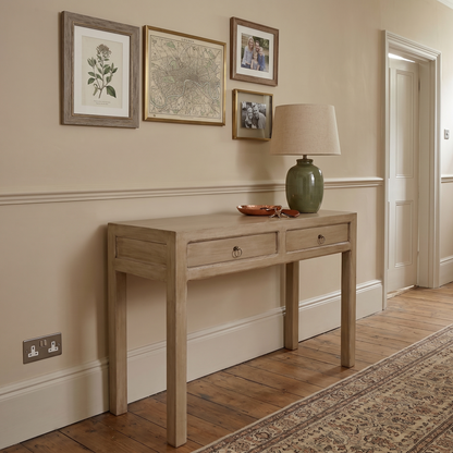 A light natural wood console table with two drawers and small round metal knobs, styled in a traditional hallway with a green ceramic table lamp, framed botanical prints, and a patterned area rug.