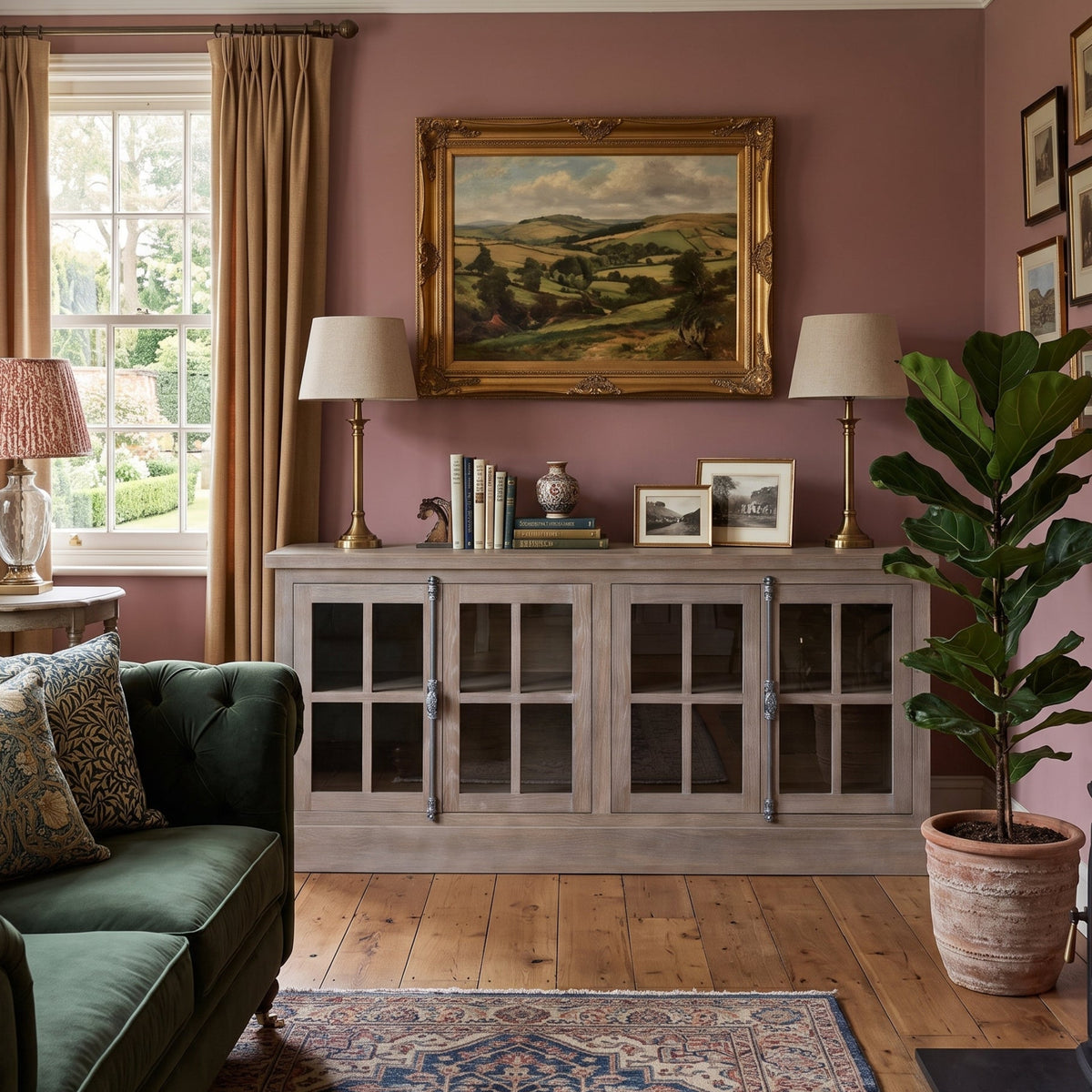 A styled living room interior featuring a dusty rose wall, a large gold-framed landscape painting, a light gray glass-front cabinet, a green sofa, a potted fiddle-leaf fig plant, and a wood-burning stove.