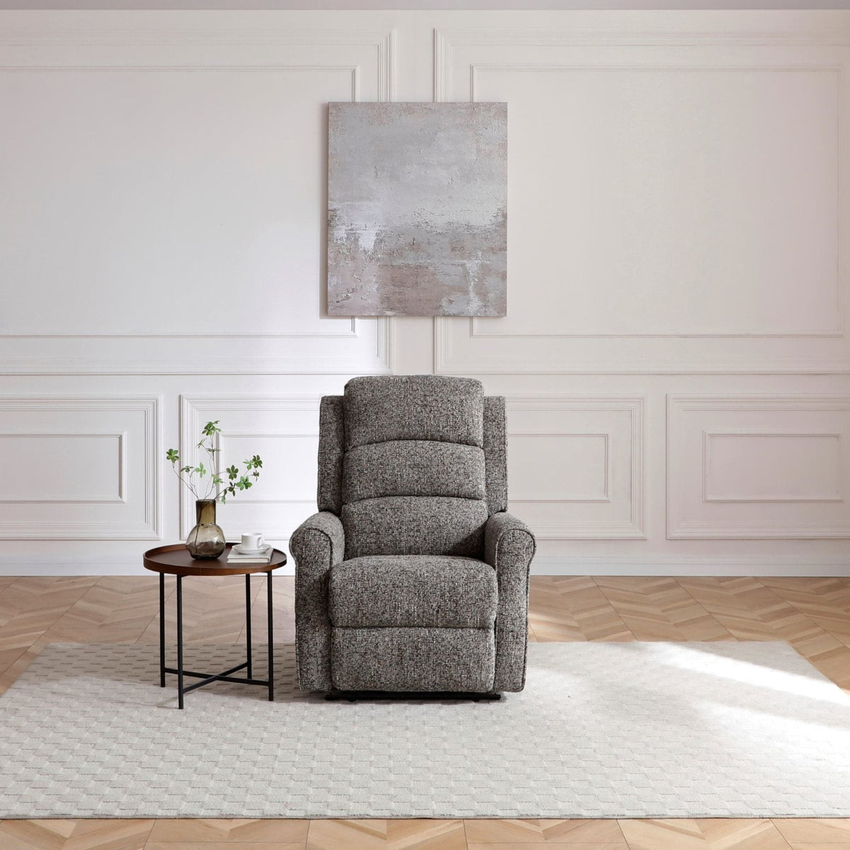 A grey upholstered recliner chair sits on a white area rug in a bright room with white paneled walls, a small round side table, and a light wood herringbone floor.